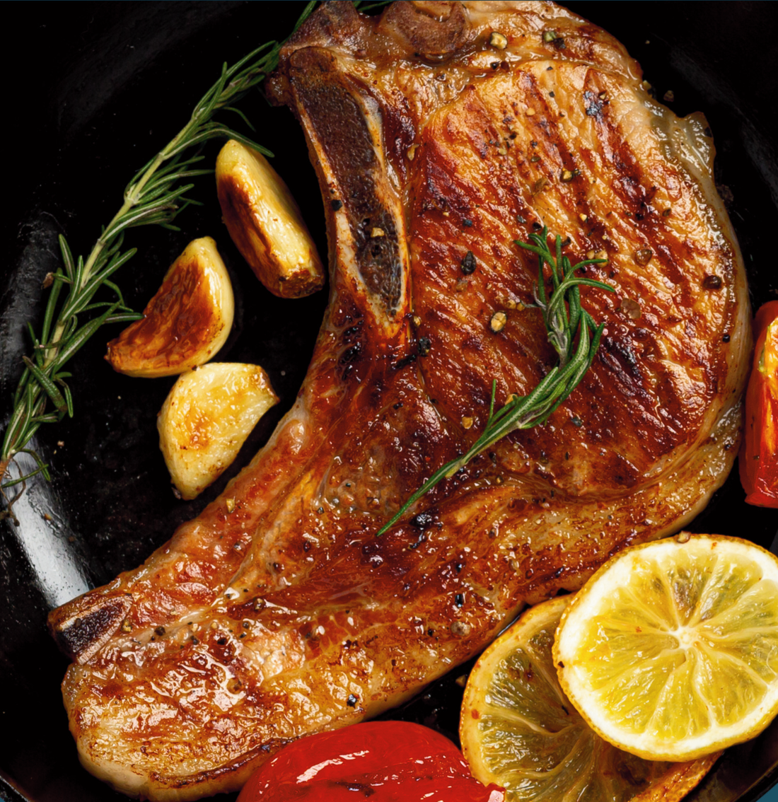 A golden-brown, cooked bone-in pork chop in a pan with a garnish of garlic, herbs, and lemon slices.