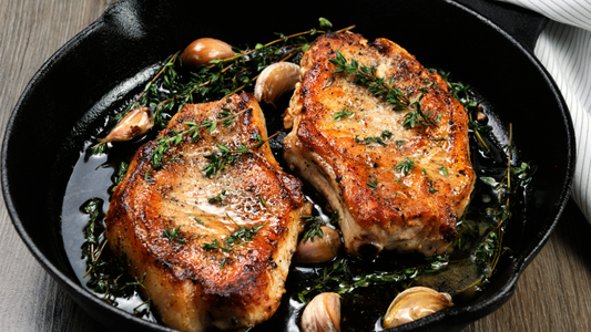 A golden-brown, cooked bone-in pork chop in a pan with a garnish of garlic, herbs, and lemon slices.