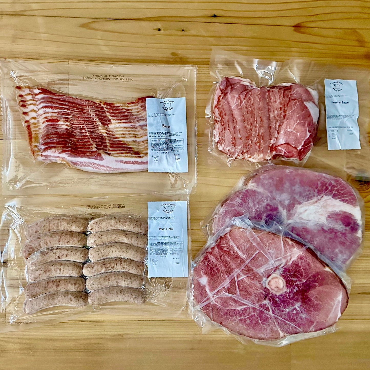 Contents of the Brunch Box from Fischer Family Farms arranged on a table. Contains 1 package bacon, 1 package Canadian bacon, 1 package pork breakfast sausage links, and 2 individually-packaged ham steaks.