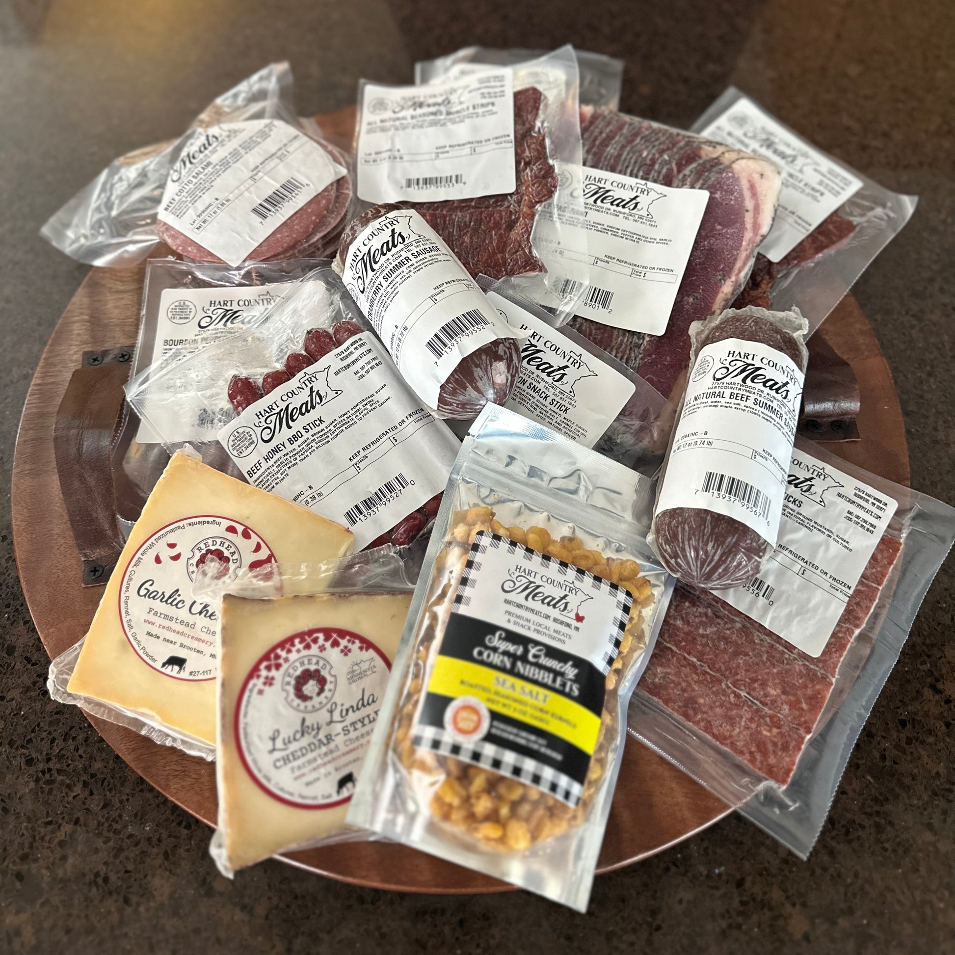 The contents of The Whole Charcuterie Box from Hart Country Meats, including 1 all natural original flavor summer sausage, 1 all natural cranberry summer sausage, 2 packs of flat strips, 2 packs of beef sticks, 2 blocks of cheese, 1 package of corn nibs, 1 package of roast beef, and 1 package of cotto salami.