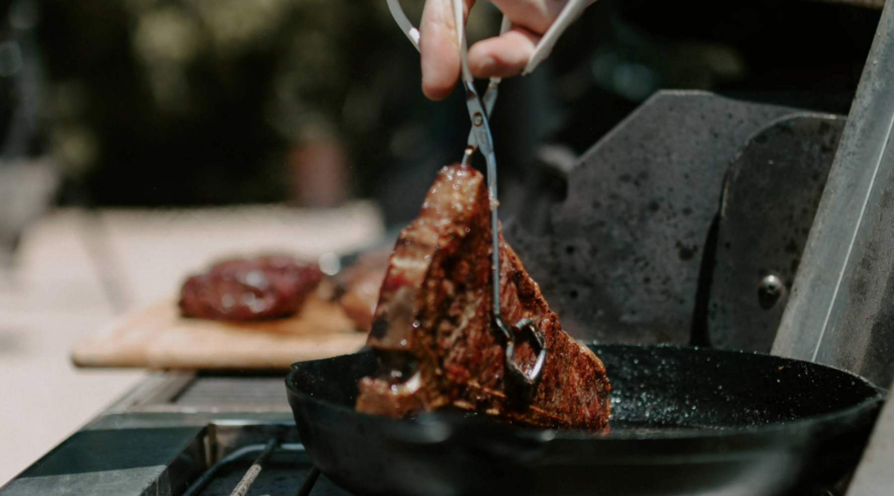 A MTRY & CO crew member placing a seared t-bone steak into a hot cast iron pan on a girll.